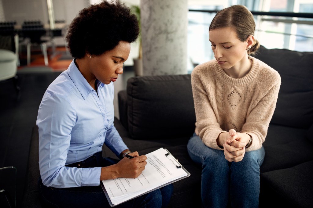 Two people sitting on a couch looking down at a clipboard. One person is writing on the clipboard.
