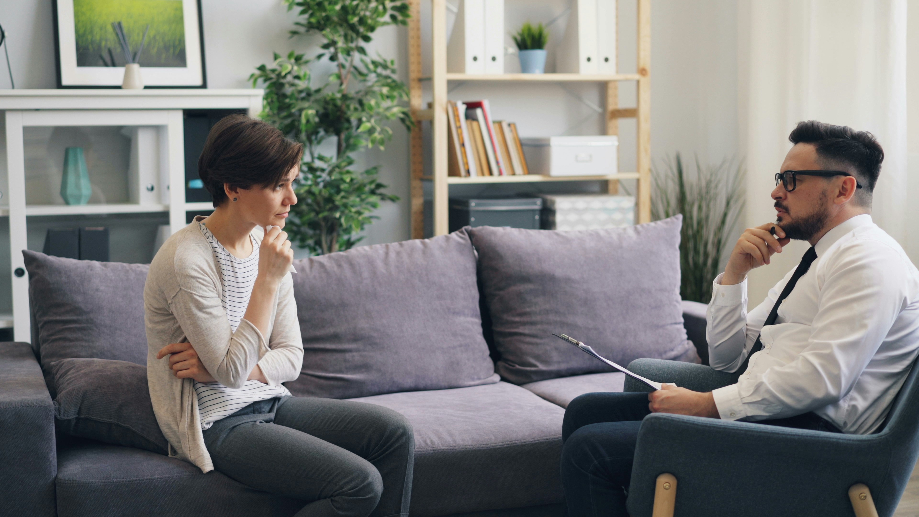 Person sitting on a couch thinking sitting across from another person sitting in a chair with a clipboard. 