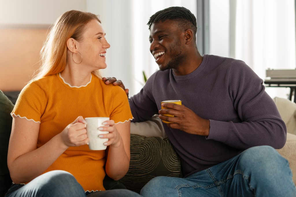 Image of a couple sitting on a couch smiling at each other. Each person is holding a mug.